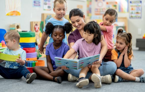 A Kindergarten teacher sits with a small group of children as she reads them a story.  They are dressed comfortably and are listening attentively.